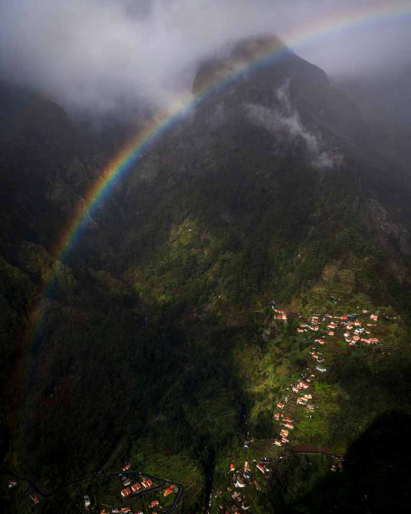 Looking down a valley to a row of houses on a cliff illuminated by a rainbow in madeira